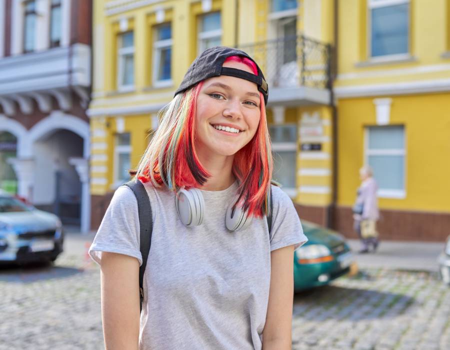 Smiling girl with colourful hair wearing a grey t-shirt and a black cap on backwards, with headphones around her neck